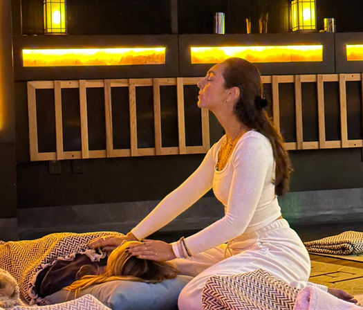 Practitioner in white giving a calming head massage to a person lying on cushions and patterned blankets in a warmly lit indoor wellness studio.