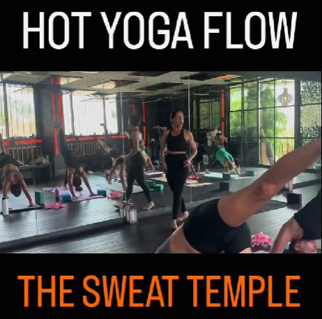 Hot yoga class in a mirrored studio — instructor leading a flowing sequence as students practice downward dog and side plank on mats with blocks in a bright, modern room.