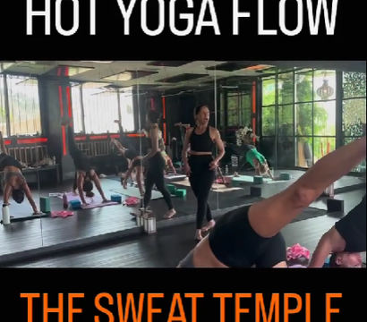Hot yoga class in a mirrored studio — instructor leading a flowing sequence as students practice downward dog and side plank on mats with blocks in a bright, modern room.