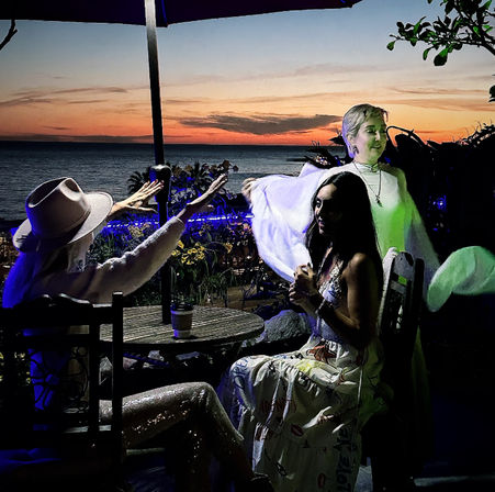 Three friends on a seaside patio at sunset — two seated at a round table with drinks under an umbrella and a third in a flowing white shawl, ocean and orange twilight in the background.
