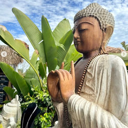 Serene wooden Buddha statue with prayer beads in a sunny tropical garden, framed by large banana leaves and a blue sky.
