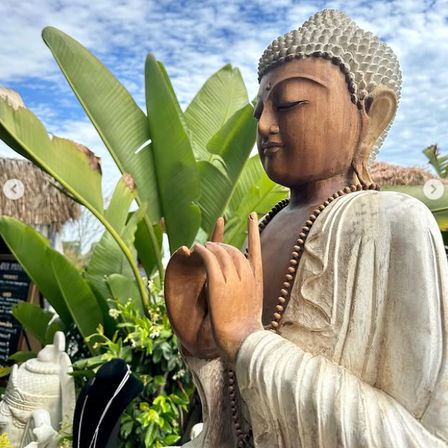 Serene wooden Buddha statue with prayer beads in a sunny tropical garden, framed by large banana leaves and a blue sky.