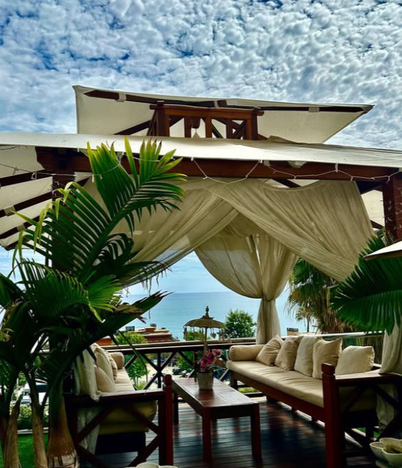 Tropical beachfront cabana with sheer drapes and cushioned wooden sofas around a coffee table, potted palms framing a turquoise ocean view under a dramatic cloudy sky.