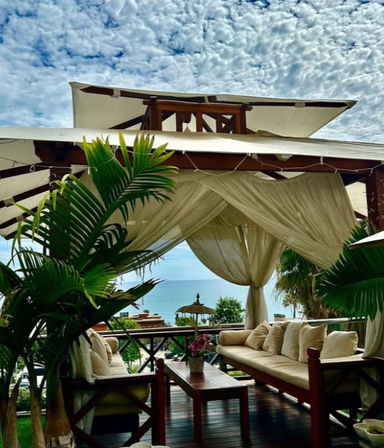 Tropical beachfront cabana with sheer drapes and cushioned wooden sofas around a coffee table, potted palms framing a turquoise ocean view under a dramatic cloudy sky.