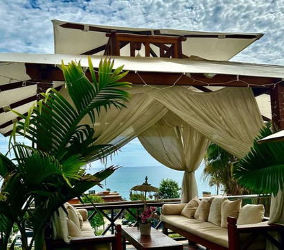 Tropical beachfront cabana with sheer drapes and cushioned wooden sofas around a coffee table, potted palms framing a turquoise ocean view under a dramatic cloudy sky.