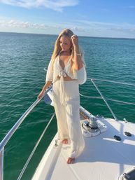 Sunlit woman in a white summer outfit standing barefoot on the bow of a yacht, holding the railing with long blonde hair blowing, turquoise sea and blue sky in the background.