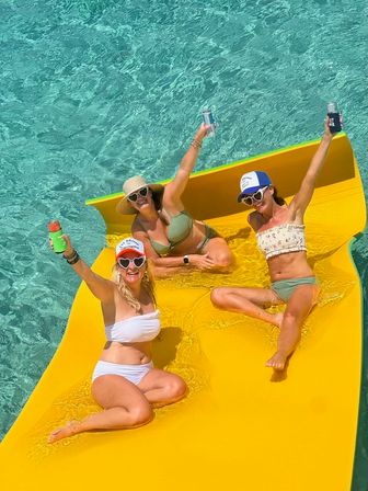Three friends relaxing on a bright yellow floating mat in clear turquoise water, wearing swimsuits, hats and heart-shaped sunglasses and raising canned drinks on a sunny tropical day