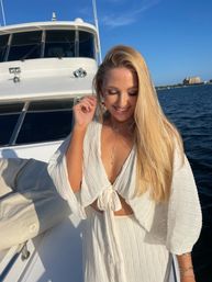 Smiling woman in a white tie-front dress on a sunlit yacht deck, long blonde hair blowing, blue ocean and distant coastal skyline under a clear sky.