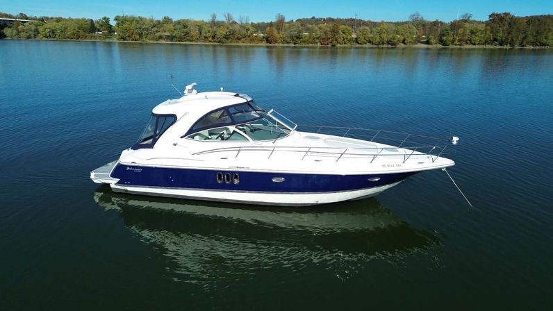 White-and-blue motor yacht anchored on calm lake water, reflecting below with a tree-lined shoreline and clear blue sky