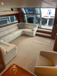 Sunlit luxury yacht salon with cream leather L-shaped sofa, zebra-print throw pillow, beige carpet, wooden table with pink headband and steps leading to aft deck dining and marina view.