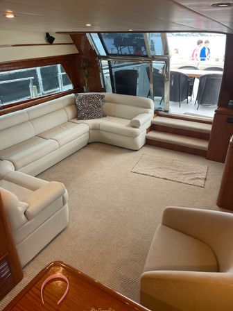 Sunlit luxury yacht salon with cream leather L-shaped sofa, zebra-print throw pillow, beige carpet, wooden table with pink headband and steps leading to aft deck dining and marina view.