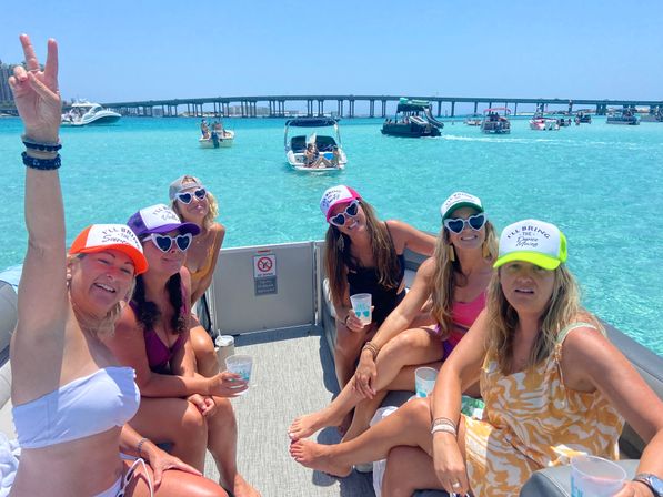 Friends on a pontoon boat enjoying a sunny boat party in turquoise coastal waters, wearing colorful caps and heart-shaped sunglasses, with other boats and a long bridge in the background.