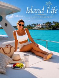 Smiling woman in sunglasses and white crop top lounging on a yacht deck over turquoise tropical water, with palm-lined waterfront villas, a straw hat, woven tote, fresh fruit plate and travel tumbler nearby.