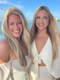 Two smiling blonde women in white summer dresses posing by the ocean under a sunny blue sky, sunlit beach vacation portrait