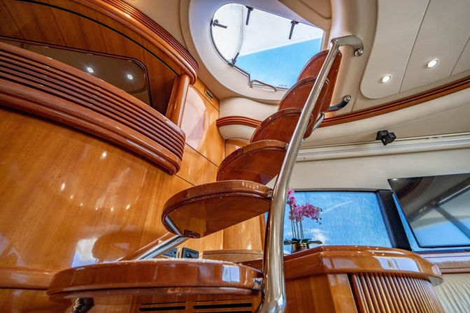 Luxury yacht interior with glossy wooden spiral staircase and stainless handrail leading up to a round hatch skylight, polished wood cabinetry, window and orchid accent.