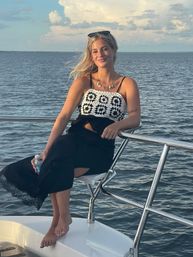 Smiling woman in a white crochet top and black skirt sitting barefoot on a yacht railing, holding a water bottle as a warm summer sunset lights calm coastal waters.