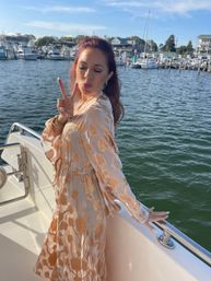 Woman in a gold-patterned white dress posing on a yacht, blowing a kiss and flashing a peace sign with a sunny marina and sailboats in the harbor behind her