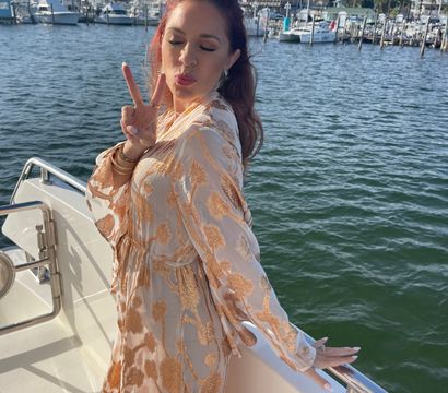 Woman in a gold-patterned white dress posing on a yacht, blowing a kiss and flashing a peace sign with a sunny marina and sailboats in the harbor behind her