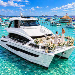 Man proposing on a flower-decorated yacht in turquoise water near a crowded sandbar with dozens of anchored boats and a thatched floating bar under a sunny blue sky