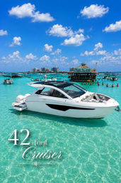 White 42-foot cruiser anchored in crystal-clear turquoise water at a busy Gulf Coast sandbar with a thatched floating tiki bar, people wading, distant high-rise skyline and blue sky dotted with puffy clouds.