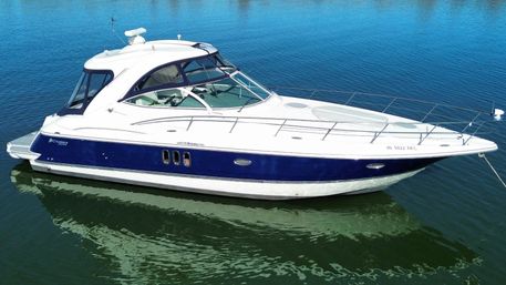 Aerial view of a sleek white-and-navy motor yacht anchored in calm blue-green water with sunlit deck and stainless-steel bow rails.