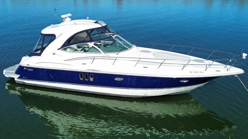 Aerial view of a sleek white-and-navy motor yacht anchored in calm blue-green water with sunlit deck and stainless-steel bow rails.