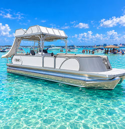White pontoon boat with canopy and slide floating on crystal-clear turquoise water near a busy sandbar with other boats and sunbathers under a bright blue sky