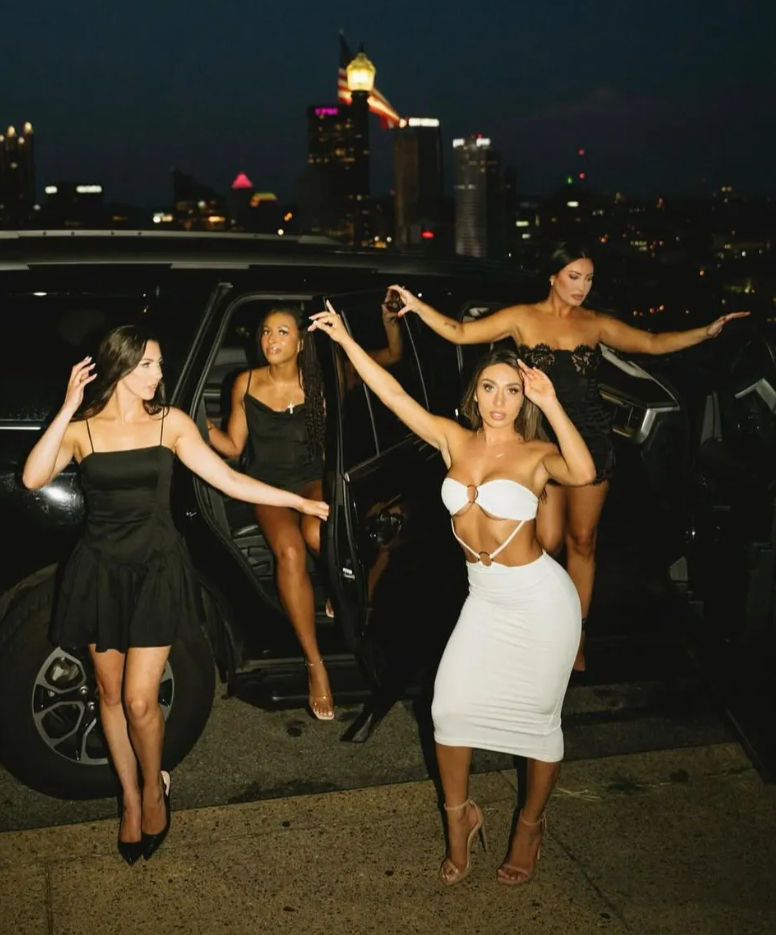 Four fashionable women stepping out of a black SUV for a glamorous night out on a downtown street with a lit city skyline in the background.