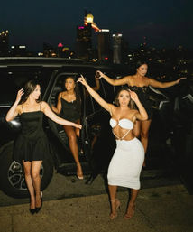 Four fashionable women stepping out of a black SUV for a glamorous night out on a downtown street with a lit city skyline in the background.