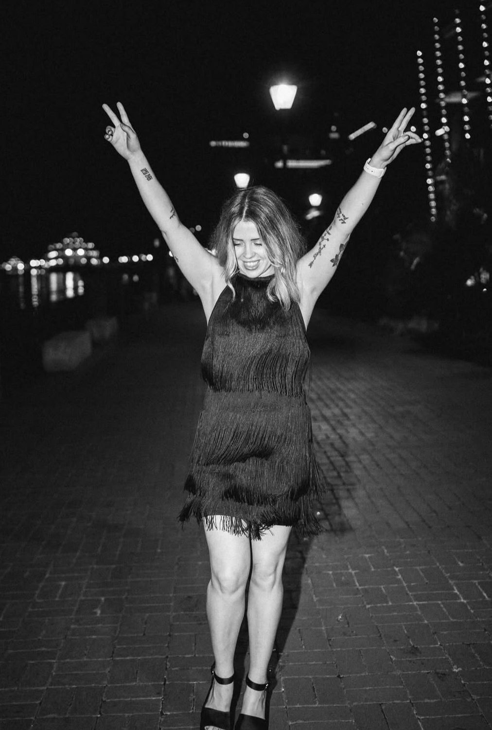 Black-and-white photo of a woman dancing on a brick waterfront promenade at night in a fringed cocktail dress, arms raised under street lamps and city lights.