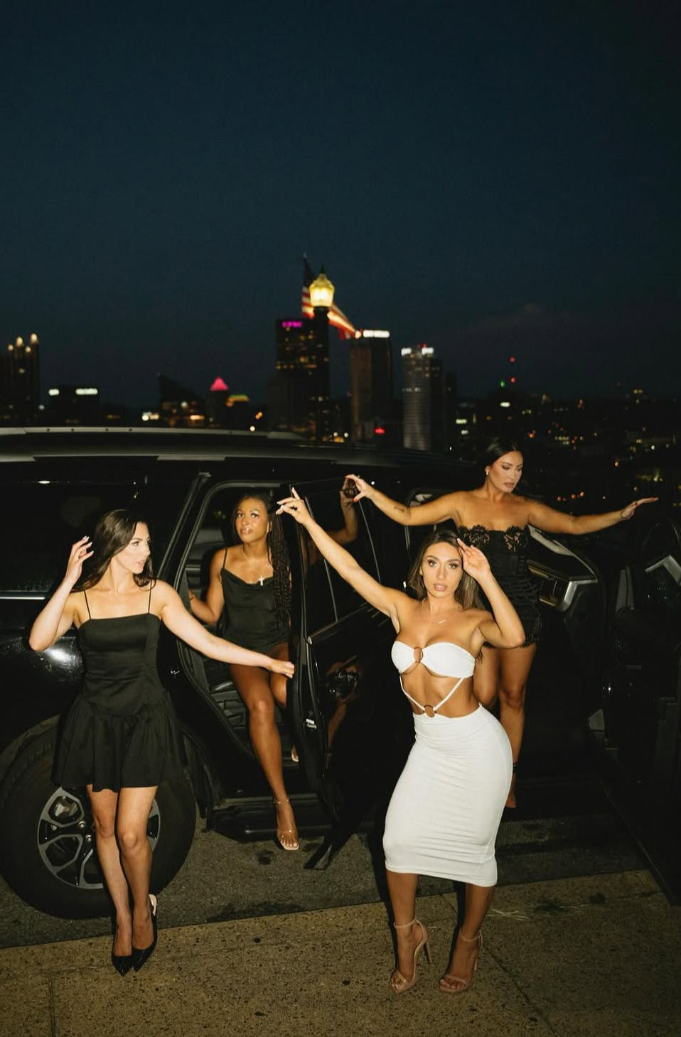 Four women in cocktail dresses stepping from a black SUV on a rooftop at night with a lit city skyline in the background — lively night-out scene.