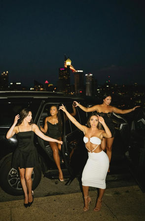 Four women in cocktail dresses stepping from a black SUV on a rooftop at night with a lit city skyline in the background — lively night-out scene.