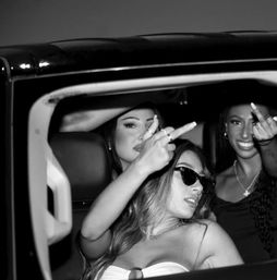 Three friends in a car at night, two playfully giving the middle finger to the camera while another wears sunglasses and a strapless top — bold nightlife vibe.