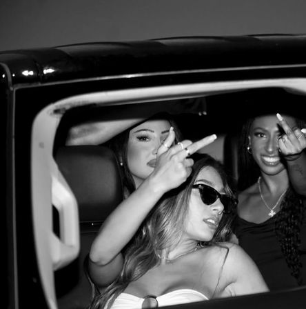 Three friends in a car at night, two playfully giving the middle finger to the camera while another wears sunglasses and a strapless top — bold nightlife vibe.