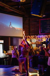 Burlesque fire performer in a sparkling costume kneeling on a neon-lit nightclub stage, blowing a vertical flame as audience members film and watch by a bar with string lights.