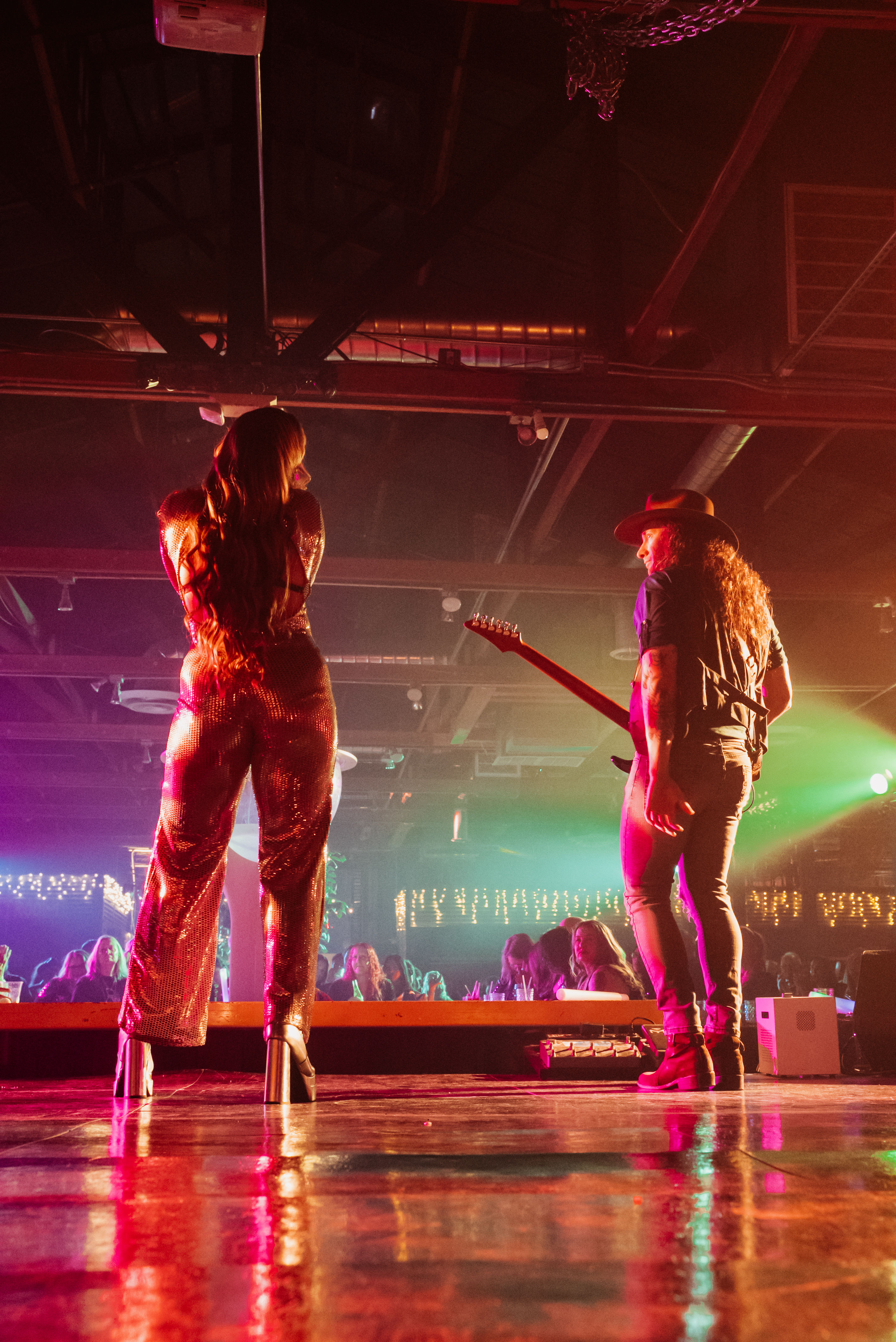 Back view of a singer in a sparkling jumpsuit and a guitarist performing on a crowded indoor live-music stage with colorful nightclub lights reflecting on the floor.