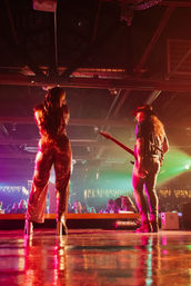 Back view of a singer in a sparkling jumpsuit and a guitarist performing on a crowded indoor live-music stage with colorful nightclub lights reflecting on the floor.