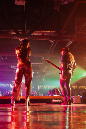Back view of a singer in a sparkling jumpsuit and a guitarist performing on a crowded indoor live-music stage with colorful nightclub lights reflecting on the floor.