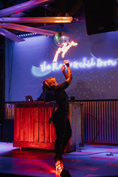 Fire performer in a sparkly outfit on an indoor nightclub stage under a disco ball, blowing a flame above a neon sign with a DJ booth and vivid purple-red lighting.
