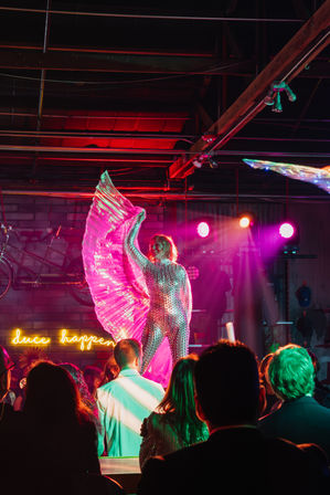 Performer in a reflective sequin bodysuit waving large pink iridescent wings on an indoor nightclub stage, lit by colorful neon and spotlights with a silhouetted audience watching.