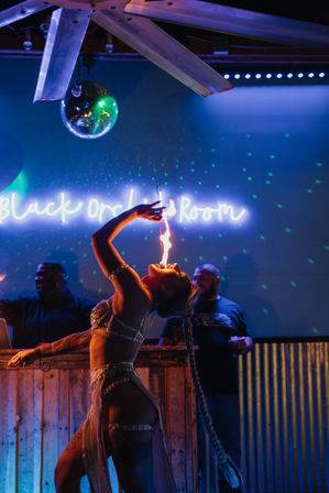 Fire-eating performer in a sparkling costume arches back on a neon-lit nightclub stage under a disco ball, breathing a vertical flame amid blue and purple party lights.