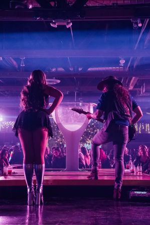 Back view of a vocalist in metallic platform boots and a hat-wearing guitarist performing on a purple-lit nightclub stage, packed crowd and drinks visible in an indoor concert venue
