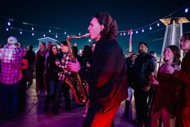 Saxophonist playing through a lively rooftop crowd under string lights at night, guests holding drinks and dancing near outdoor heaters