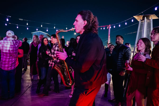 Saxophonist playing through a lively rooftop crowd under string lights at night, guests holding drinks and dancing near outdoor heaters