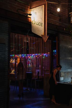 Dim urban bar entrance at night with a retro illuminated arrow sign, beaded curtain, colorful string lights, bar stools and a silhouette of a person leaning at a table.