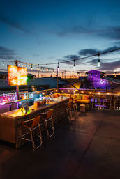 Rooftop bar at twilight with colorful string lights and neon sign, long outdoor bar with stools, cozy cushioned lounge seating and city rooftops on the horizon