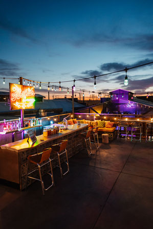 Rooftop bar at twilight with colorful string lights and neon sign, long outdoor bar with stools, cozy cushioned lounge seating and city rooftops on the horizon