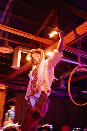 Fire performer with long braided hair twirling a flaming torch overhead on a dim industrial event stage, bathed in purple-orange lights with an aerial hoop in the background.