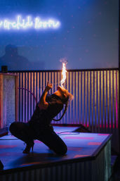 Performer in a sequined black outfit kneeling on a small stage, leaning back and breathing a tall flame upward with neon blue signage and corrugated metal backdrop lit by pink and purple stage lights.