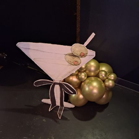 Paper martini cocktail cutout with three olive garnishes resting on a cluster of metallic gold balloons against a black backdrop — playful party centerpiece / event decor.